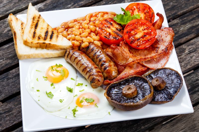 Teller mit englischem Frühstück: Bacon, Spiegelei, Würstchen, Tomaten, Champignons, Bakes Beans und Toast