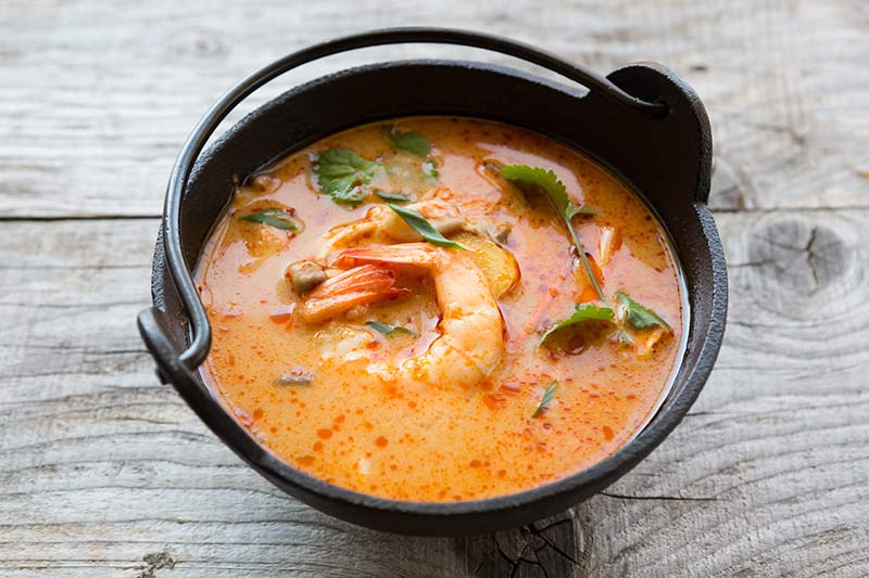 Tom Yum Kong in schwarzem Schälchen. Garnele, grünes Gewürz und orange Flüssigkeit sind zu sehen.
